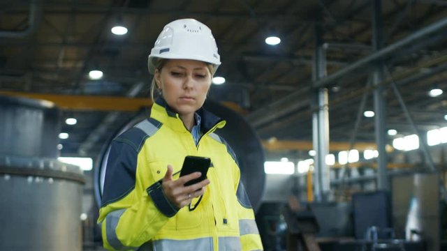 Female Industrial Worker in the Hard Hat Uses Mobile Phone While Walking Through Heavy Industry Manufacturing Factory. In the Background Various Metalwork Project Parts Lying 