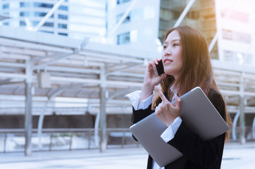 beautiful young asian businesswoman talking with smartphone and holding laptop outdoor in the city.