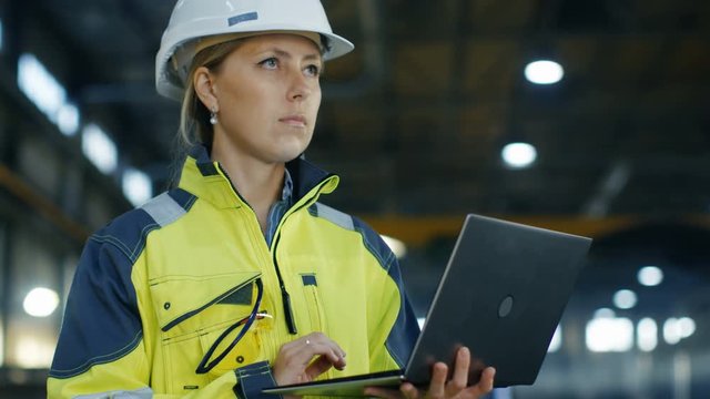 Portrait Female Industrial Engineer in the Hard Hat Uses Laptop Computer while Standing in the Heavy Industry Manufacturing Factory. In the Background Various Metalwork Project Parts Lying 