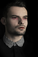 Fototapeta premium Close up face of a Vampire Man. Fashion male model as Vampire with tattoo (cross) on the neck and full lips and bleeding eyes. Portrait of pretty Vampire man looking away with serious look.