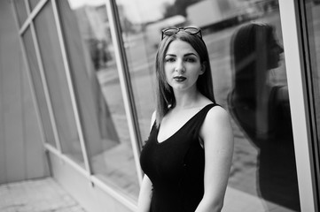 Red haired stylish girl in sunglasses wear in black, against large windows. Street fashion portrait.