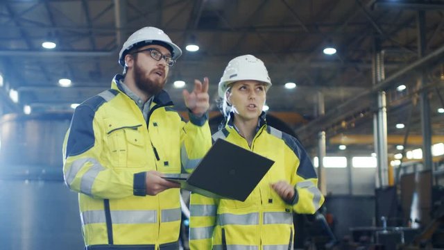 Back View Shot of Male and Female Industrial Engineers Having Discussion and Use Laptop While Walking Through Heavy Industry Manufacturing Factory. They Wear Hard Hats and Safety Jackets.