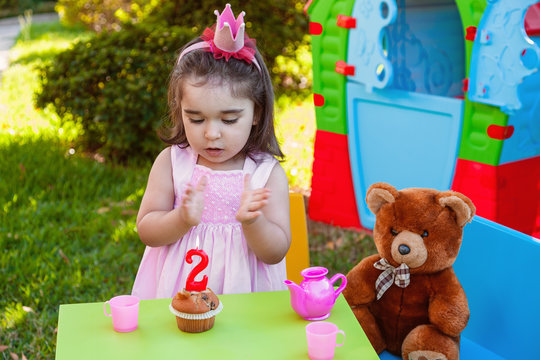 Baby Toddler Girl In Outdoor Second Birthday Party Clapping Hands At Cake With Teddy Bear As Best Friend, Playhouse And Tea Set. Pink Dress And Crown.