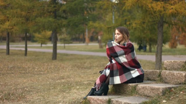 Girl in the autumn park.
Slow motion. A young girl sits on a rock in the park and drinks a hot drink.