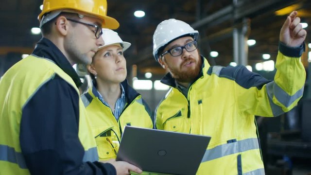 Three  Industrial Engineers Talk with Factory Worker while Using Laptop. They Work at the Heavy Industry Manufacturing Facility.