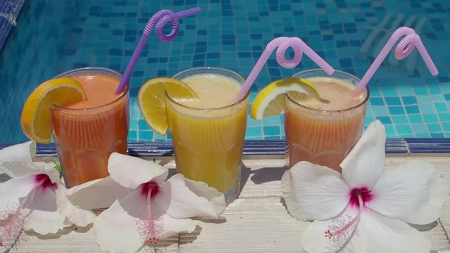 Three glasses of tropical fruit juice with a drinking straw on the side of the pool on a hot day