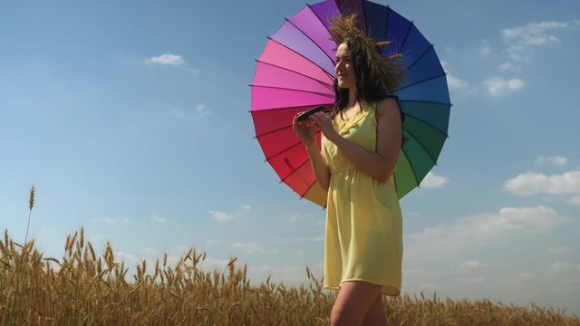 Woman in yellow dress walks on wheatfield rotating rainbow umbrella and smiling