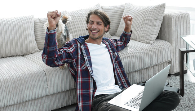 happy guy rejoicing and raising his hands sitting near the sofa in the living room.