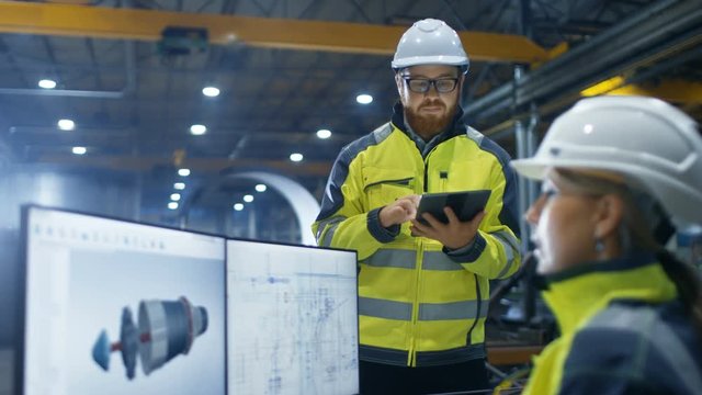 Inside the Heavy Industry Factory Female Industrial Engineer Works on Personal Computer She Designs 3D Engine Model, Her Male Colleague Talks with Her and Uses Tablet Computer. - Powered by Adobe