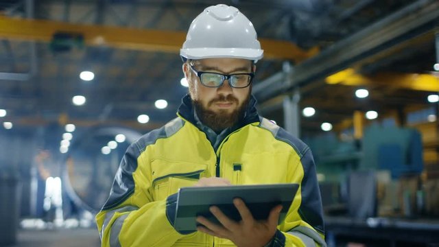 Industrial Engineer in Hard Hat Wearing Safety Jacket Uses Touchscreen Tablet Computer. He Works at the Heavy Industry Manufacturing Factory.