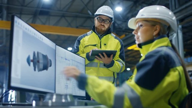 Inside the Heavy Industry Factory Female Industrial Engineer Works on Personal Computer She Designs 3D Engine Model, Her Male Colleague Talks with Her and Uses Tablet Computer. Low Angle Footage. - Powered by Adobe