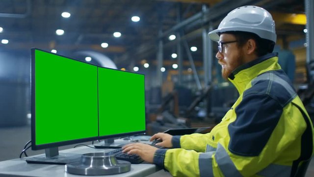 Inside the Heavy Industry Factory Industrial Engineer Works on the Personal Computer with Two  Green Mock-up Screens.