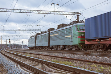 Obraz premium Lokomotywa pociągu towarowego ciągnie skład kontenerów na peronach torów kolejowych.