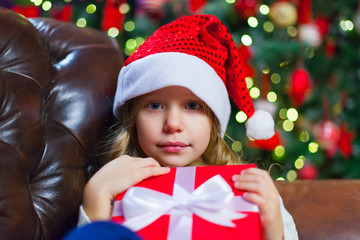 Little girl sitting by the tree holding a Christmas gift In the 