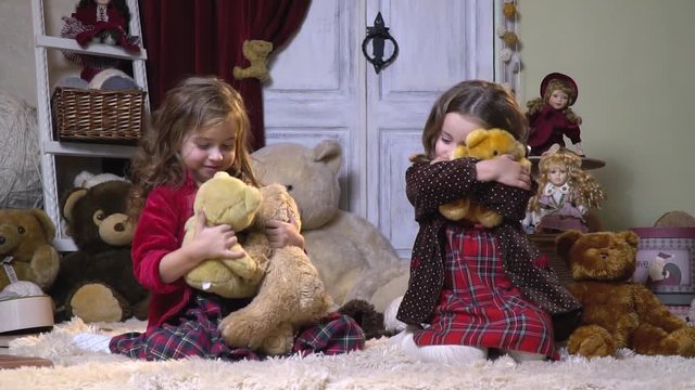 Two girls in dresses hug plush toys sitting on a nap carpet, slow motion