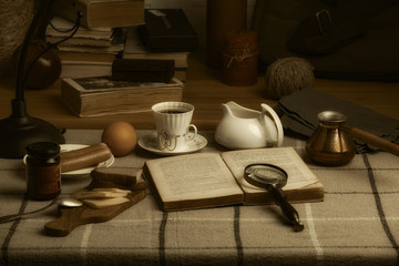 An open book, a cup of coffee, a magnifier and food on the table. A bag, books, a candle and other things on the windowsill.