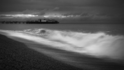 Long Exposure Black & White Image of Palace Pier, Brighton, UK with Wave Breaking Against the Shore in the Foreground 