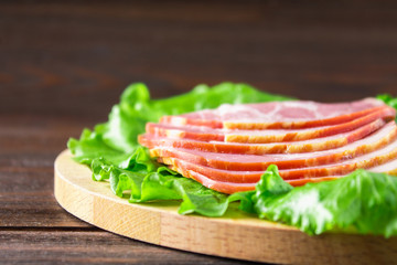 Sliced ham with fresh green lettuce leaves on a round cutting board. Meat products on a brown wooden table.