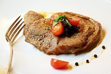 Closeup image of fresh juicy roasted lamb chop on white plate with fork, meat sauce, black peppercorns, fresh tomatoes and herbs