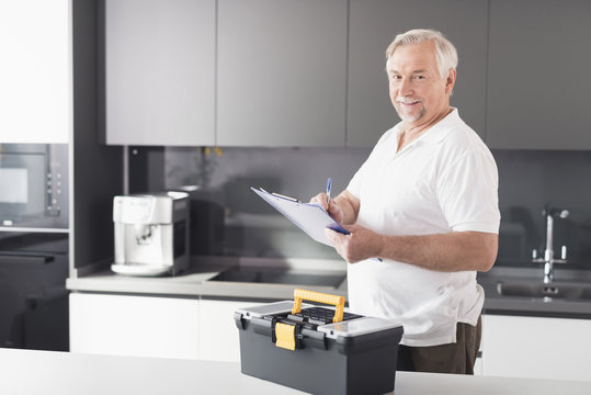 The man is in the kitchen. He holds a form for papers in his hands. Next to him stands a black box for the instrument.