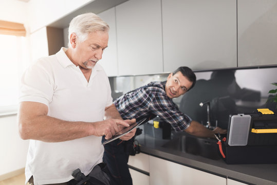 Two plumbers are standing in the kitchen. An elderly man looks something on his tablet while young man repairs the tap.