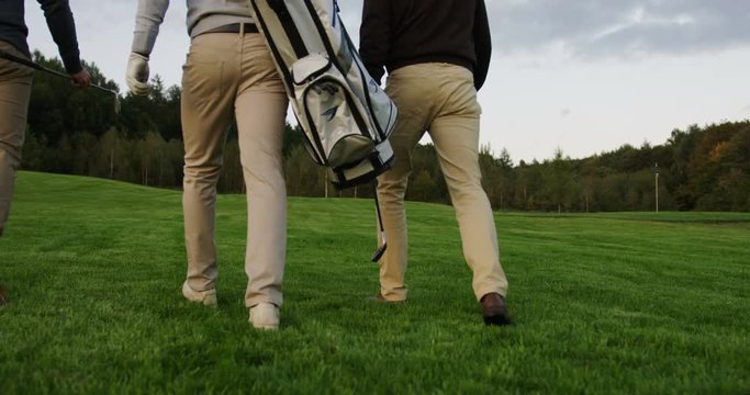 Camera follows three men who going to play golf with their equipment on the green field. From behind and below.