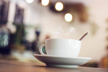White cup with steaming hot coffee on table with bokeh background at coffee shop
