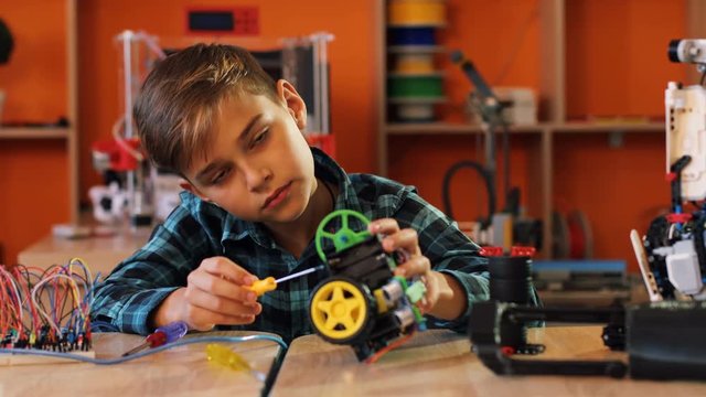 Portrait shot of nice blonde boy with the screwdriver assembling a robot and smiling straight into the camera in the modern comfortable playing space of classroom.