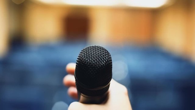 Hand holding microphone closeup view empty blurred conference hall background rehearsal repetition public speech speaker orator lector expressive speaking unrecognisable nobody indoors media urban