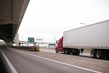 Red big rig semi truck with long reefer trailer transporting frozen cargo by interstate highway with overpass bridge