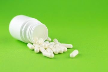 Close up white pill bottle with spilled out capsules on green background with copy space. Focus on foreground, soft bokeh. Pharmacy drugstore concept