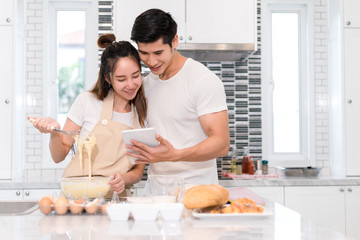 Obraz na płótnie Canvas Couple making bakery in kitchen room, Young asian man and woman together cooking cake and bread with egg, looking menu from tablet in the flour happy relaxing in at home