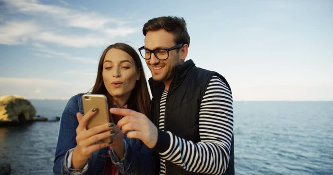 Young attractive woman and man watching photos at the phone screen on the beautiful seacoast background. Outdoors
