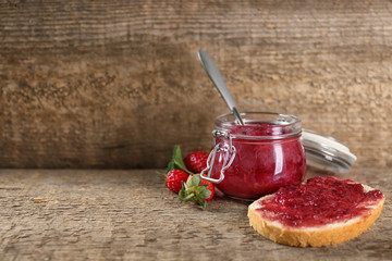 Piece of bread and strawberry jam in jar on wooden table