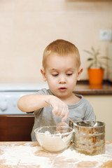 Little kid boy helps mother to cook Christmas ginger biscuit in light kitchen. Happy fair-haired child in gray T-shirt 2-3 years at the table with iron sieve and flour in weekend morning at home.