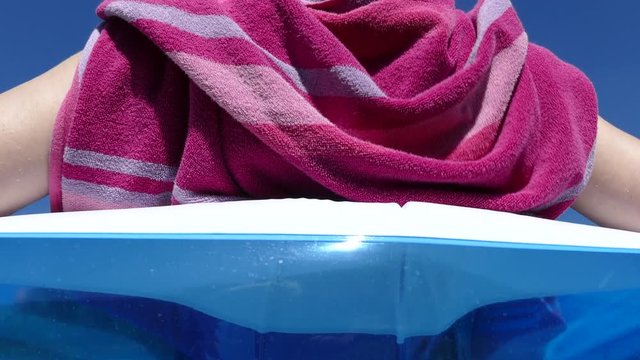 Upward dolly shot of a young woman relaxing in an inflatable garden pool against a clear blue sky, taking a towel off her shoulders and turning sideways.
