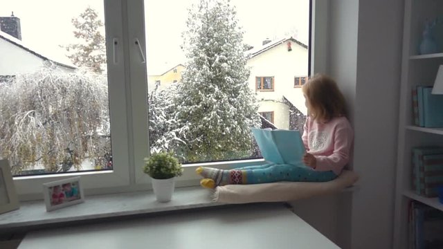 Slow motion. Portrait of a pretty little girl reading at the window. It is snowfall