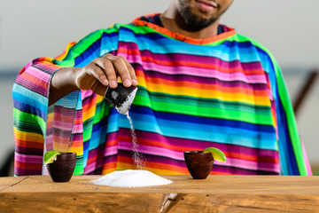 Mexican gold tequila. Selective focus on salt on the table, on the background Mexican bartender in colorful poncho. Concept of preparation tequila