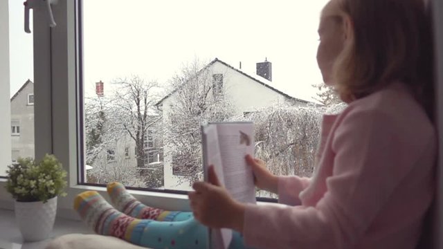 Slow motion. Portrait of a pretty little girl reading at the window. It is snowfall