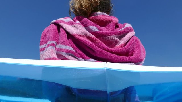 Upward dolly shot of a young woman relaxing in an inflatable garden pool against a clear blue sky, with a towel around her shoulders.