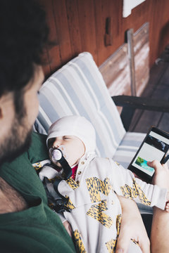 Father with sleeping baby boy using mobile phone while sitting on chair