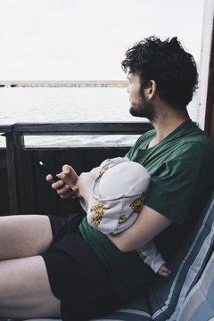 Thoughtful father with baby boy holding mobile phone while sitting on chair by lake