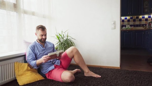 Young happy bearded man sitting on floor at home and using tablet