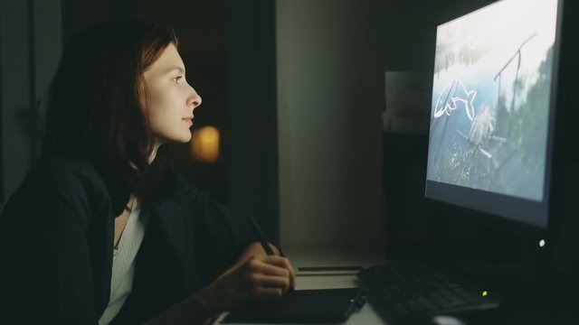 Concentrated young woman designer working in office at night using computer and graphics tablet to finish job