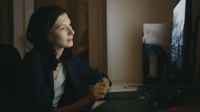 Concentrated woman designer working in office at night using computer with graphics tablet and laptop