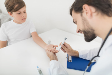 The doctor takes a blood sample from the boy to check it for sugar. The boy patiently endures the procedure