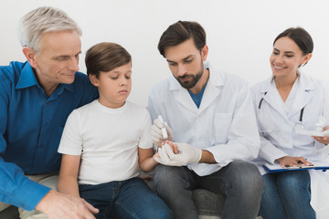 A boy with a grandfather in a medical clinic. The doctor takes a blood sample from the boy with a scarifier.