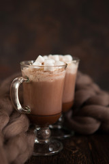Two glasses with Hot chocolate garnished with whipped cream, marsmallow and cocoa powder on  wooden background with Cozy plaid from merinose wool
