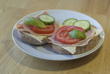 close up fresh home made rye bread sandwich with ham cheese sliced cucumbre tomatoes and basil leaves on white plate on wooden table