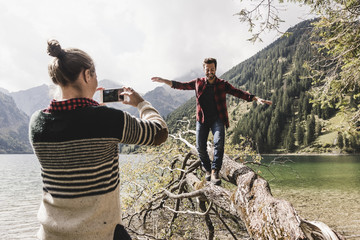 Austria, Tyrol, Alps, woman taking cell phone picture of man balancing on tree trunk at mountain lake